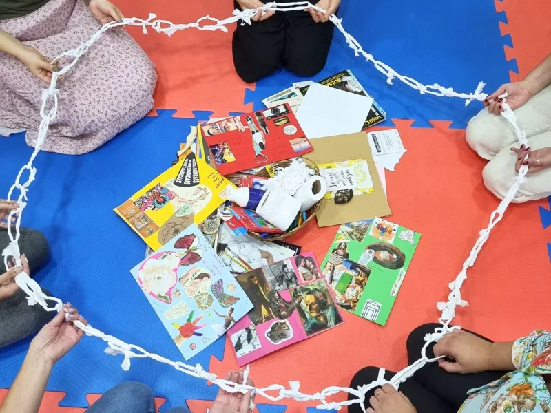 Mulheres sentadas em círculo no chão, segurando uma corrente de papel feita à mão, com colagens e materiais criativos no centro da roda