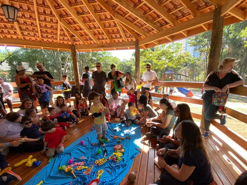 Criança em pé sobre o pano azul com instrumentos de brinquedo, cercada pelas famílias durante a musicalização